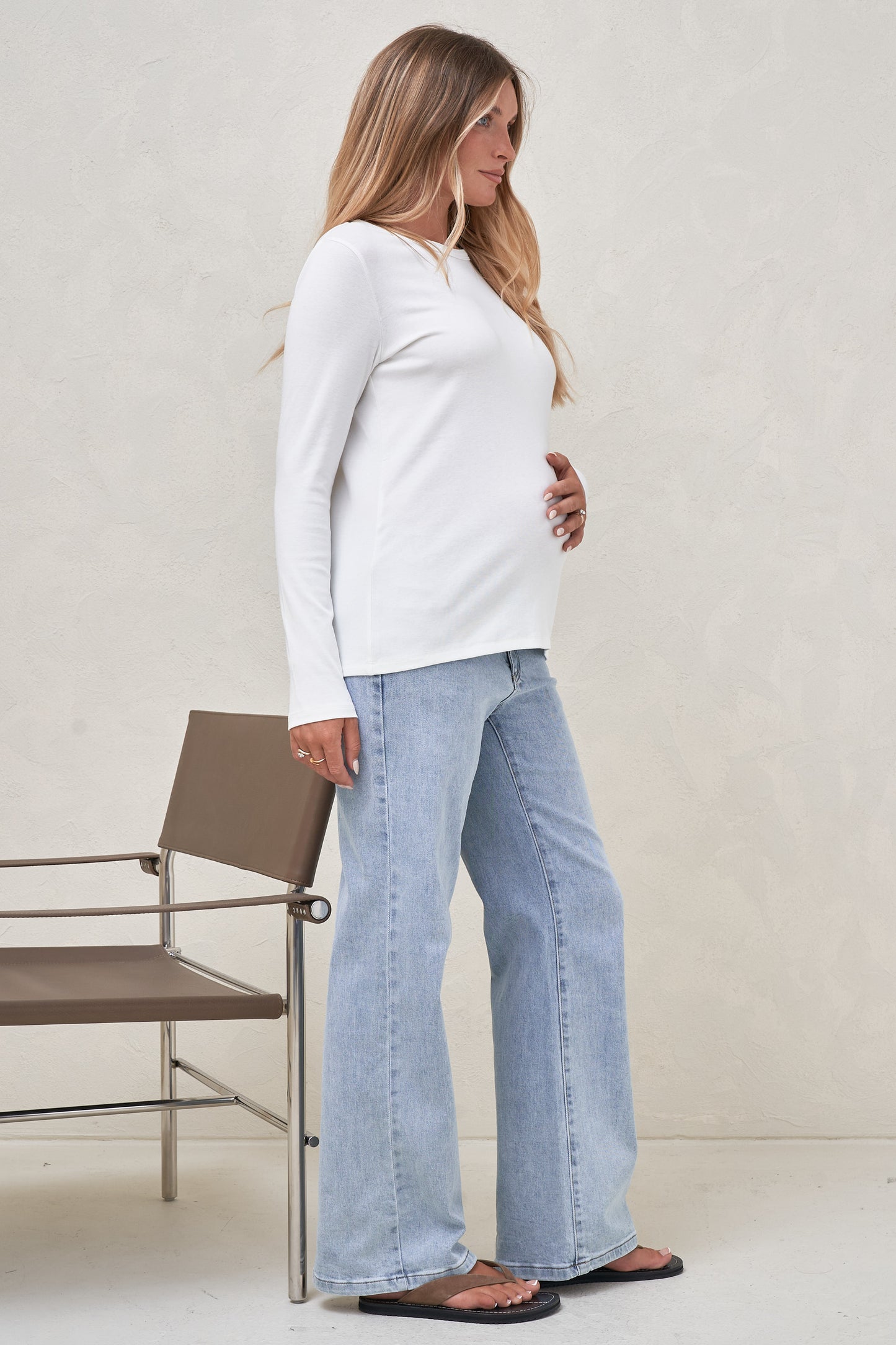 Side view of pregnant model wearing a white long sleeve top worn out over light blue maternity jeans
