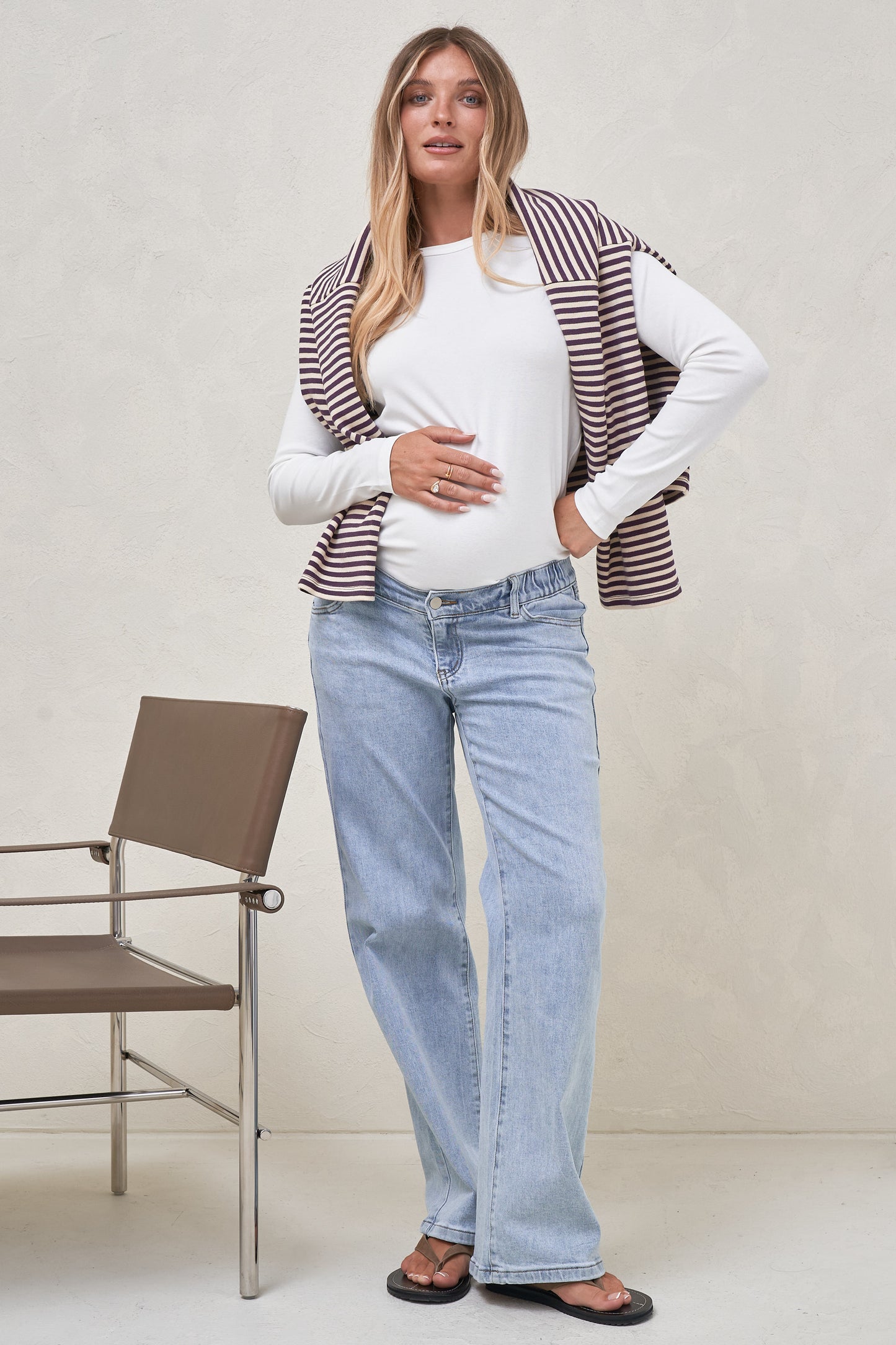 Pregnant model wearing a white long sleeve top with light blue maternity jeans and a stripe top draper over her shoulders